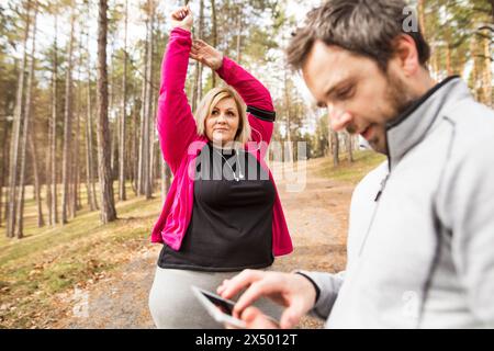 Übergewichtige Frau streckt sich vor dem Laufen, Personal Trainer überprüft ihr Profil, Leistung auf dem Tablet. Sport im Freien für Menschen mit Adipositas Stockfoto