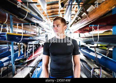 Portrat eines jungen Kanufahrers, der mitten in gestapelten Kanus steht. Das Konzept des Kanufahrens als dynamischer und abenteuerlicher Sport. Stockfoto