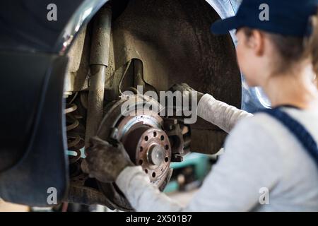 Weibliche Automechanikerin, die das Auto auf dem Aufzug hebt und die Pausen überprüft. Schöne Frau, die in einer Garage arbeitet und blaue Overalls trägt. Stockfoto