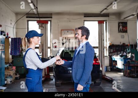 Eine Automechanikerin spricht mit dem Kunden und übergibt die Schlüssel des reparierten Autos. Schöne Frau, die in einer Garage arbeitet und blaue Overalls trägt. Stockfoto