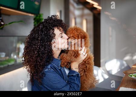 Porträt einer schönen lockigen brünetten Frau, die ihr Pudeltier in einem Restaurant kuschelt. Elegante Frau mit ihrem kleinen Hund genießt das Essen im Innenbereich. Stockfoto
