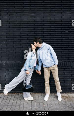 Junger Mann und asiatische Frau küssen sich an einer schwarzen Ziegelmauer. Ein Paar küsst sich an einer farbigen Wand. Stockfoto