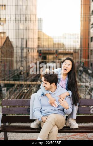 Interrassisches Paar, das auf einer Bank sitzt und in einer urbanen Landschaft rumhängt. Junge Mann und Frau sitzen auf einer Bank über einer Eisenbahn auf der Straße der Stadt. Stockfoto