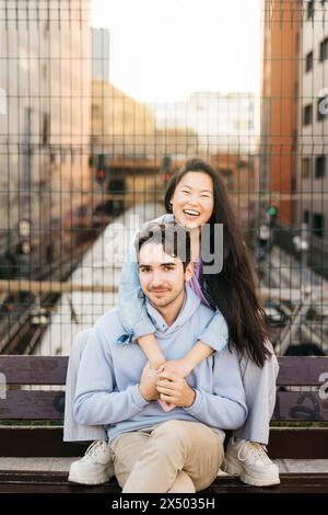 Interrassisches Paar, das auf einer Bank sitzt und in einer urbanen Landschaft rumhängt. Junge Mann und Frau sitzen auf einer Bank über einer Eisenbahn auf der Straße der Stadt. Stockfoto