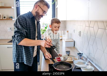 Junge, der Vater hilft, Pfannkuchen zu machen. Vater verbringt Zeit mit dem Sohn zu Hause, macht zusammen Snacks, kocht. Vatertagskonzept. Stockfoto