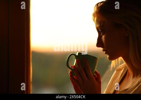 Seitenporträt einer melancholischen Frau, die Kaffee trinkt und bei Sonnenuntergang durch ein Fenster schaut Stockfoto
