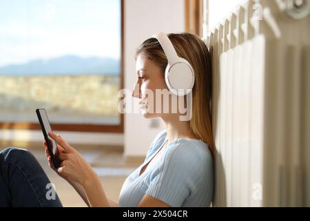 Profil einer seriösen Frau, die sich am Heizkörper erhitzt und Musik mit Kopfhörern und Mobiltelefon zu Hause hört Stockfoto