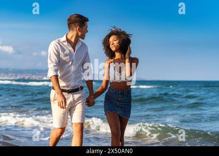 Junge interrassische Paare - gehen Hand in Hand an einem Sandstrand, genießen einen romantischen und fröhlichen Spaziergang zusammen - warm, lebhaft. Stockfoto
