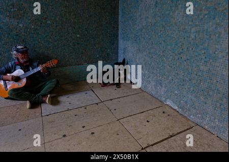 Ein älterer Mann schnappt sich seine Gitarre, um Spenden zu erhalten, sitzt in einer Unterführung, und sein schwarzer Hund liegt in der Nähe. Stockfoto