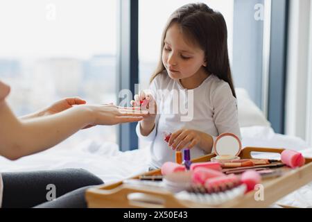 Glückliche Mutter und Tochter sitzen zu Hause im Bett und machen Maniküre, kümmern sich um ihren Körper. Das süße kleine Mädchen lernt, ihre Nägel zu malen, indem es aufträgt Stockfoto