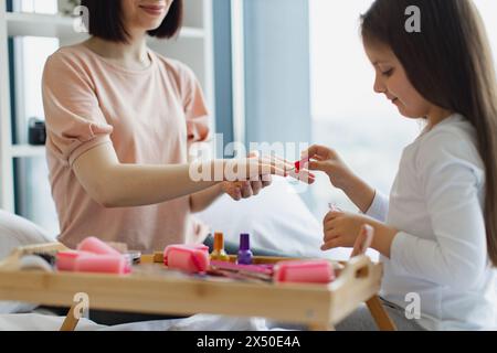 Glückliche Mutter und Tochter sitzen zu Hause im Bett und machen Maniküre, kümmern sich um ihren Körper. Das süße kleine Mädchen lernt, ihre Nägel zu malen, indem es aufträgt Stockfoto