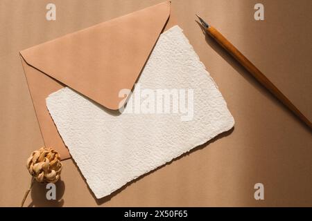 Blick von oben auf ein leeres Stück hausgemachter Deckelkantenkarte, Umschlag, Blume und Kalligrafie-Stift auf einem Holztisch Stockfoto