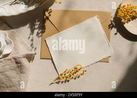 Blick von oben auf ein leeres Stück hausgemachte Deckelkantenkarte und Umschlag auf einem Tisch mit Mimosa-Blumen und Geschirr Stockfoto