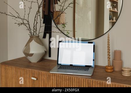 Öffnen Sie den Laptop mit einem leeren Bildschirm auf einem Sideboard neben einer Vase und einer Kerze Stockfoto
