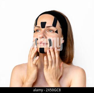 Weibliches Schönheitsmodell, das im Studio kinesio Gesichtsbänder auf ihrem Gesicht vor weißem Hintergrund aufträgt. Gesichtskonturierung und Hautstraffung. Stockfoto