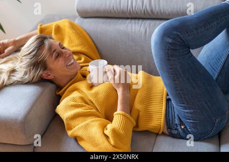 Reife Frau, die sich zu Hause auf der Couch mit einer Kaffeetasse entspannt, lächelnd und fröhlich aussah Stockfoto