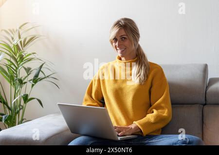 Lächelnde junge Frau, die auf einem Sofa sitzt und einen Laptop benutzt, mit einer Zimmerpflanze neben ihr Stockfoto