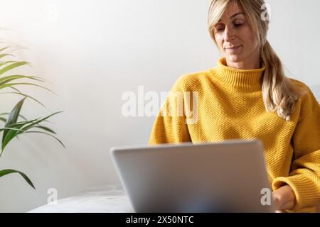 Fokussierte Frau in gelbem Pullover mit Laptop auf Sofa, mit einer Zimmerpflanze im Hintergrund Stockfoto