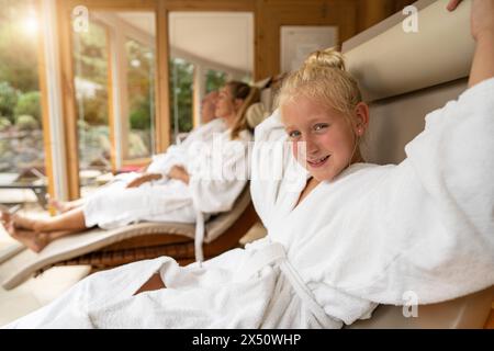 Ein junges Mädchen lächelt im Vordergrund und ihre Familie entspannt sich in Bademänteln im Hintergrund in einer hellen Spa-Hotellounge Stockfoto