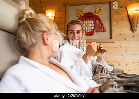Die Mutter, die eine Teetasse hält, spricht mit ihrer Tochter, während sie sich in einer Liege in einem Wellness-Spa-Hotel entspannt, beide lächeln Stockfoto