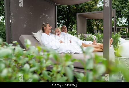 Eine fröhliche Familie von drei Personen, Mutter, Vater und Tochter, lacht zusammen, während sie sich in einer luxuriösen Spa-Cabana im Freien entspannen Stockfoto