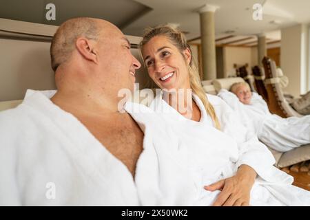 Ein Paar teilt sich einen freudigen Moment in einer Spa-Lounge, wobei ihre Tochter im Hintergrund sichtbar ist, alle in weißen Bademänteln Stockfoto