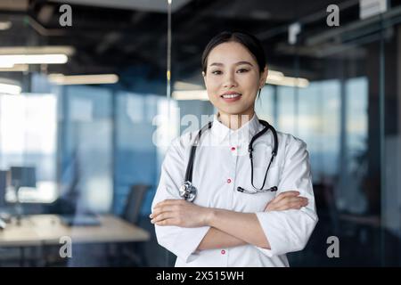 Eine kompetente und einladende junge Ärztin in einem Labormantel mit überkreuzten Armen und einem Stethoskop, die selbstbewusst in einem Büro steht. Stockfoto