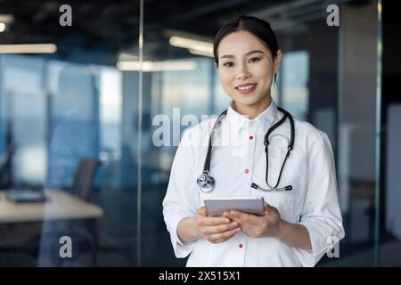 Eine professionelle asiatische Ärztin mit Stethoskop steht selbstbewusst in einem modernen Büro und hält ein digitales Tablet, das Gesundheit und Technologie widerspiegelt. Stockfoto