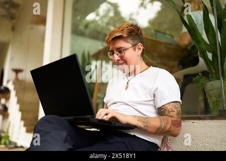 Tätowierte Gen Z Transgender Person arbeitet auf einem Laptop in einem gemütlichen Büro und zeigt einen zwanglosen, inklusiven Arbeitsbereich. Konzentriert und dennoch komfortabel, repräsentiert Stockfoto