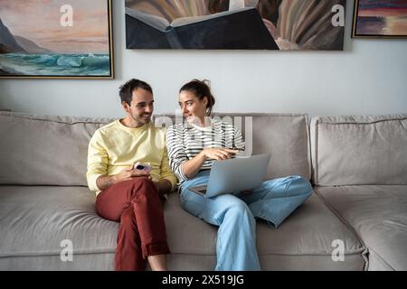 Die interessierte Frau zeigt einen glücklichen Mann auf dem Laptop-Bildschirm, sitzt auf der Couch, redet, kauft zu Hause online. Stockfoto