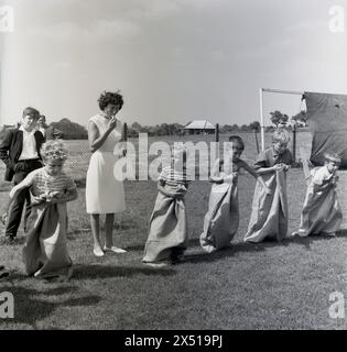 1964, historisch, draußen auf einem Feld, Junge, die für den Beginn des Kindersack-Rennens gesäumt sind, auf deinen Spuren, bereit... Go, England, Großbritannien. Ein Sackrennen ist ein beliebtes Sommerspiel, bei dem die Teilnehmer mit ihren Beinen im Sack bis zur Taille das Sackmaterial in ihren Händen sammeln und dann in Richtung Ziellinie springen oder hüpfen. Stockfoto