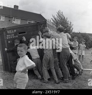 1964, historisch, draußen auf einem Feld, eine Gruppe von Kindern, die eine große Holz-/Metall-Fracht-Kiste auf die Seite schieben, um zu spielen und Spaß zu haben. Stockfoto