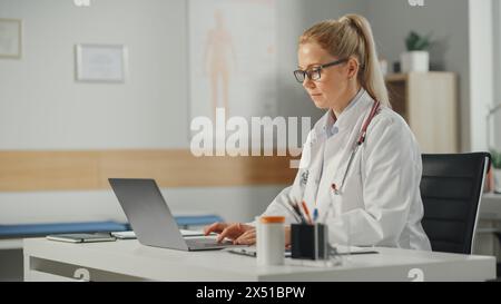 Erfahrene Ärztin mit weißem Mantel, die in ihrem Büro an einem Laptop arbeitet. Kaukasischer medizinischer Arzt in Gläsern arbeitet mit Testergebnissen, Patienten-Behandlungsplanung. Stockfoto