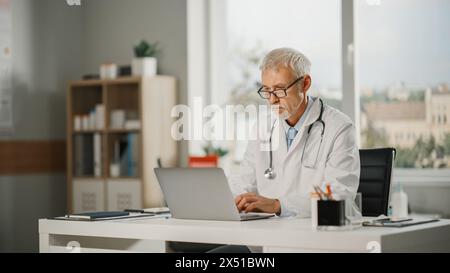 Porträt eines erfahrenen männlichen Arztes mittleren Alters mit weißem Mantel, der in seinem Büro an einem Laptop arbeitet. Senior Medical Care Professional Working. Stockfoto
