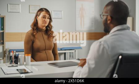 Afroamerikanischer Familienarzt spricht mit jungen schwangeren Patienten während der Konsultation in einer Gesundheitsklinik. Erfahrener Arzt im Labormantel, der hinter einem Tisch mit Laptop im Krankenhausbüro sitzt. Stockfoto