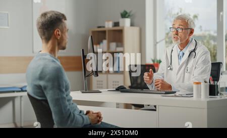 Familienarzt mittleren Alters spricht mit jungen männlichen Patienten während der Konsultation in einer Gesundheitsklinik. Oberarzt im Labormantel, der hinter einem Computerpult im Krankenhausbüro sitzt. Stockfoto