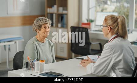 Die weibliche Hausärztin sitzt hinter einem Schreibtisch und spricht während der Konsultation in einer Gesundheitsklinik mit einer Seniorin. Arzt im Labormantel verschreibt Medikamente an ältere Patienten in der Krankenhauspraxis. Stockfoto