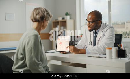 Afroamerikanischer Familienarzt zeigt Herzanalyseergebnisse auf einem Tablet-Computer einer älteren Patientin während der Konsultation in einer Gesundheitsklinik. Arzt sitzt hinter einem Schreibtisch im Krankenhausbüro. Stockfoto
