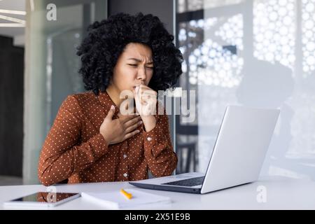 Eine junge afroamerikanische Frau, die sich unwohl fühlt, Husten, während sie an ihrem Schreibtisch arbeitet. Das Bild zeigt ihr Unwohlsein und das Gesundheitskonzept am Arbeitsplatz. Stockfoto