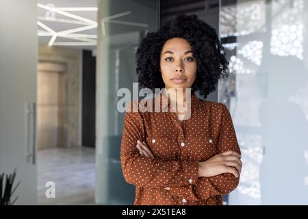 Eine junge afroamerikanische Frau steht selbstbewusst in einer Büroumgebung, trägt ein stylisches Polka Dot-Shirt und zeigt einen subtilen, durchdachten Ausdruck. Stockfoto