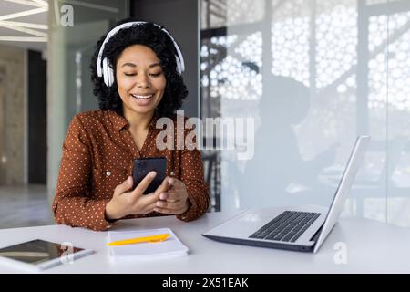 Eine fröhliche Afroamerikanerin nutzt ihr Smartphone, während sie Musik über Kopfhörer an ihrem modernen Arbeitsplatz hört. Sie zeigt einen freudigen Ausdruck. Stockfoto