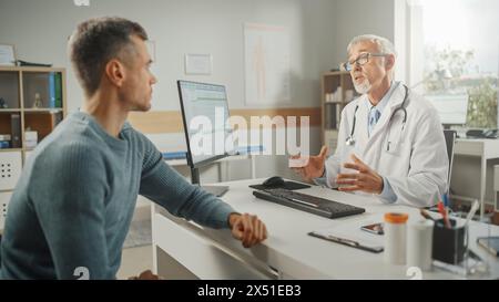 Familienarzt mittleren Alters spricht mit jungen männlichen Patienten während der Konsultation in einer Gesundheitsklinik. Oberarzt im Labormantel, der hinter einem Computerpult im Krankenhausbüro sitzt. Stockfoto