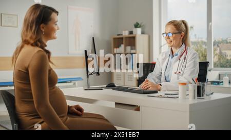 Weibliche Hausärztin spricht mit jungen schwangeren Patienten während der Konsultation in einer Gesundheitsklinik. Erfahrener Arzt im Labormantel, der hinter einem Computerpult im Krankenhausbüro sitzt. Stockfoto