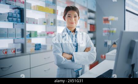 Apotheke Drugstore: Porträt der schönen asiatischen Apothekerin mit weißem Labormantel, Blick auf Kamera, Lächeln, hinter ihrem Checkout Counter, Regale mit Medizin-Paketen, Gesundheitsprodukte Stockfoto
