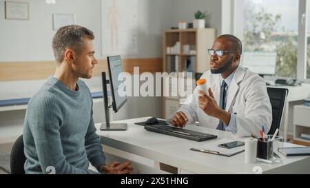 Afroamerikanischer Familienarzt spricht mit Young Male Patient während der Konsultation in einer Gesundheitsklinik. Schwarzer Arzt im Labormantel und Brille, der hinter einem Computerpult im Krankenhausbüro sitzt. Stockfoto