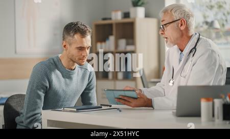 Erfahrener Hausarzt mittleren Alters zeigt Analyseergebnisse auf einem Tablet-Computer an männlichen Patienten während der Konsultation in einer Gesundheitsklinik. Arzt sitzt hinter einem Schreibtisch im Krankenhausbüro. Stockfoto