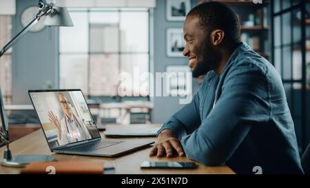 Schöner afroamerikanischer Mann, der einen Videoanruf mit weiblicher Ärztin auf Laptop-Computer hat, während er hinter dem Schreibtisch im Wohnzimmer sitzt. Freiberuflicher Mitarbeiter, der von zu Hause aus arbeitet und über das Internet einen Medic spricht. Stockfoto