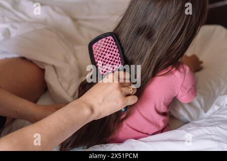 Mom kämmt das Haar ihrer Tochter mit einem Kamm, während sie im Bett sitzt Stockfoto