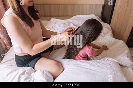 Mom kämmt die Haare ihrer Tochter mit Kamm, während sie im Bett sitzt Stockfoto