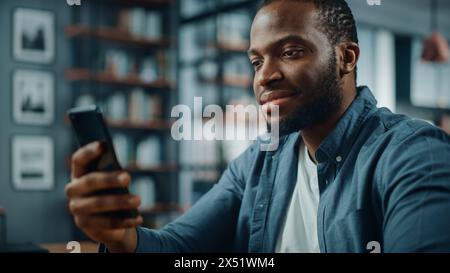 Glücklicher schwarzer Afroamerikaner, der das Smartphone benutzt, während er am Tisch im Wohnzimmer sitzt. Aufgeregter Mann, der zu Hause lächelt und mit Kollegen, Kunden und Freunden über das Internet spricht. Stockfoto
